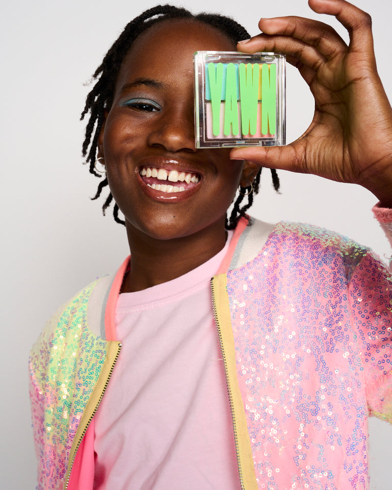 Photo of a girl smiling and holding a YAWN eyeshadow palette in front of her left eye.
