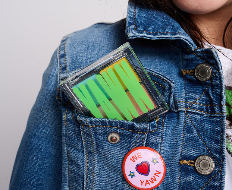 Close up photo of a girl wearing a denim jacket with YAWN eyeshadow palette peeking out of her jacket pocket. She is also wearing a button that says WE LOVE YAWN.